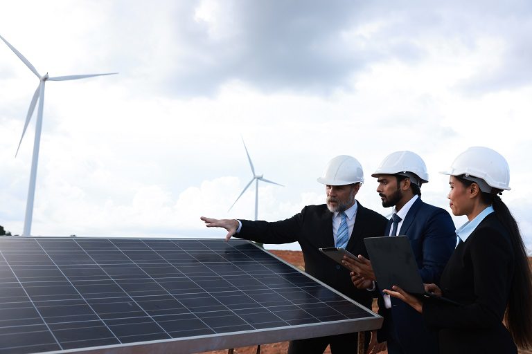 Two engineers inspecting solar panels with wind turbines in the background.