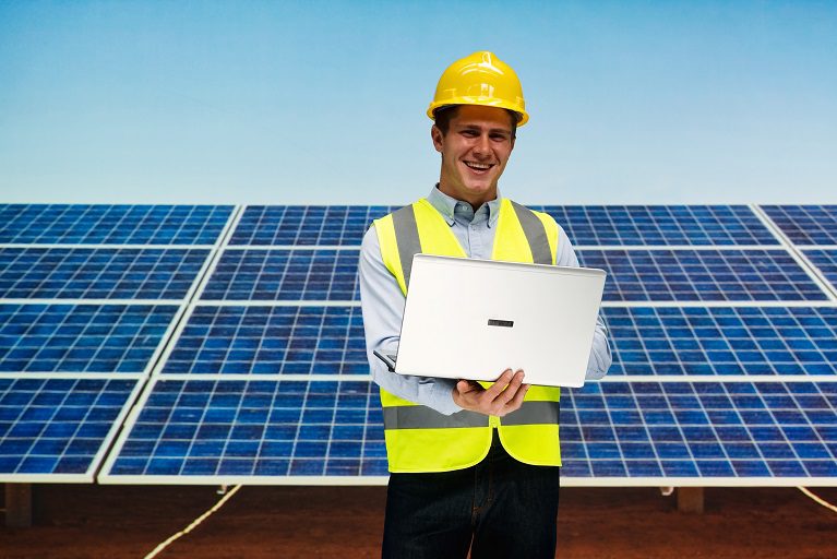Engineer in safety gear working on solar panel installation with laptop.