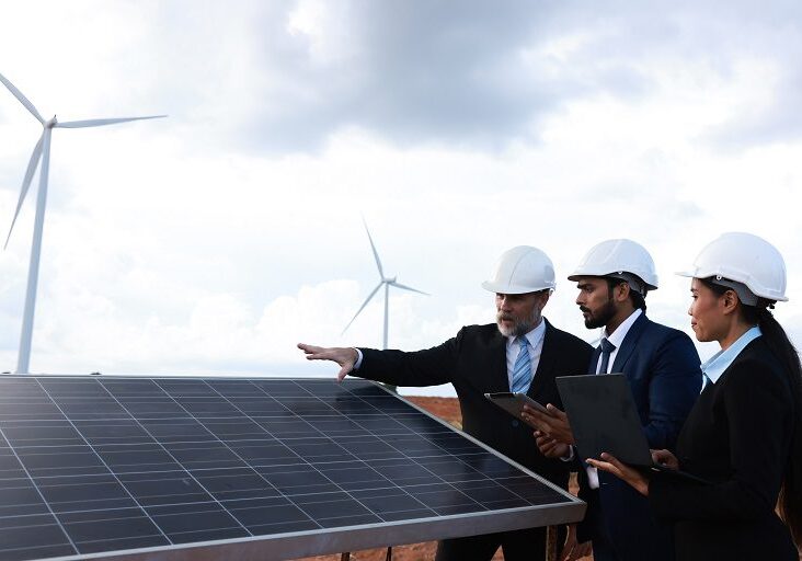 Two engineers inspecting solar panels with wind turbines in the background.