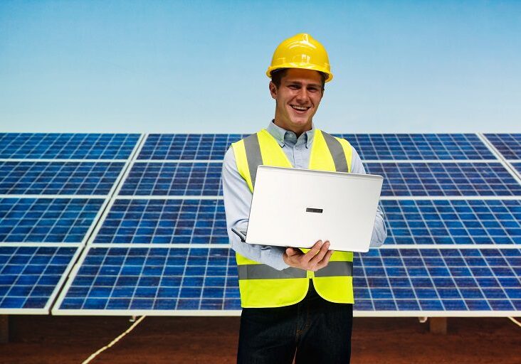 Engineer in safety gear working on solar panel installation with laptop.